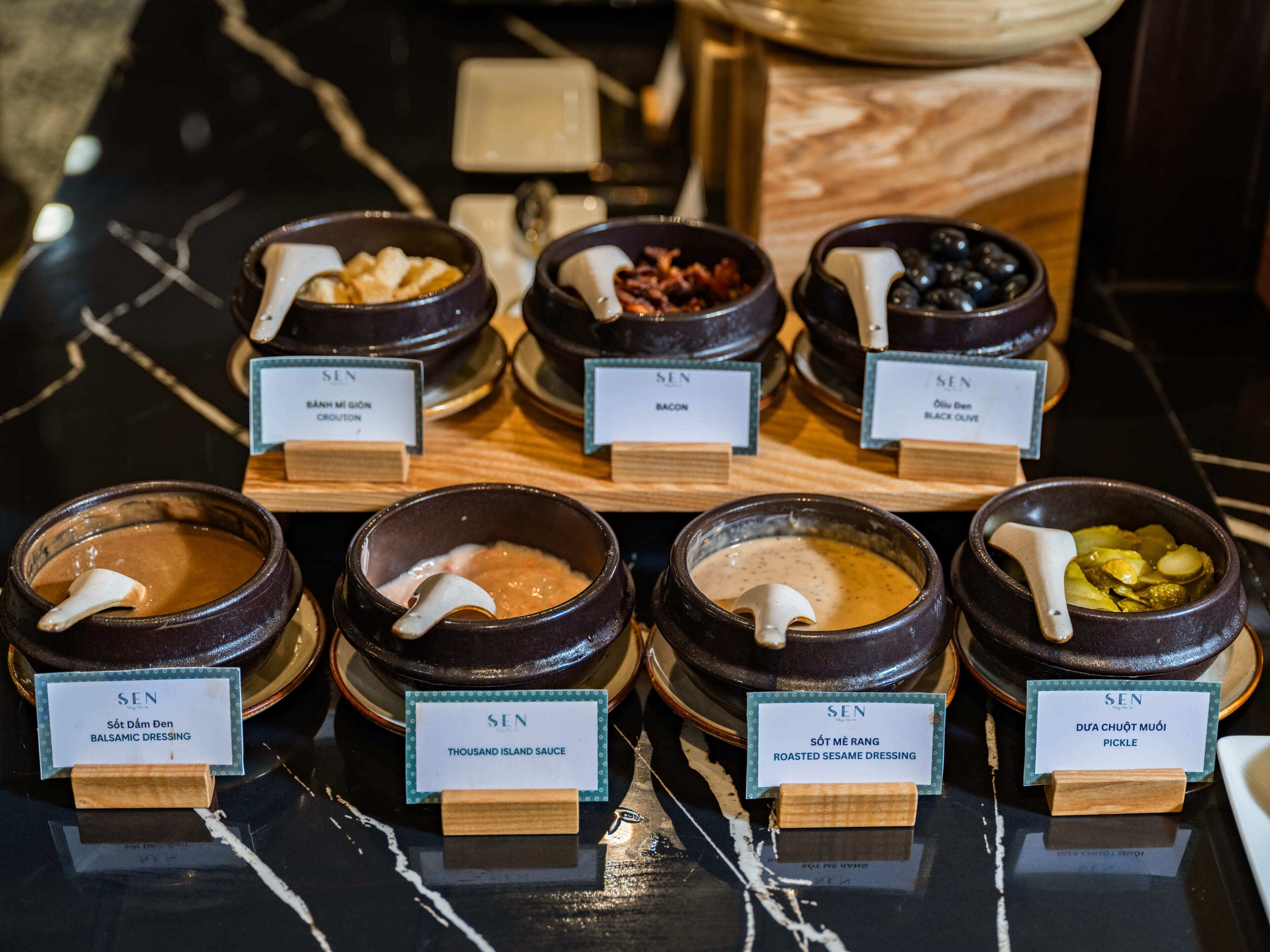 A selection of condiments in ceramic bowls with labels, arranged on a wooden board.