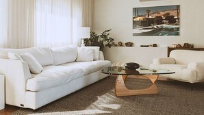 A modern living room with a white sofa, a glass coffee table, and a painting of a pool on the wall.