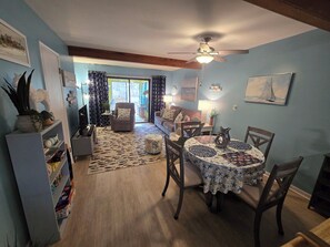 A cozy living room with a dining area, featuring a ceiling fan, a sofa, a coffee table, and a bookshelf. - Misty Mare Townhouse - Gorgeous home on Chincoteague Island (Chincoteague)