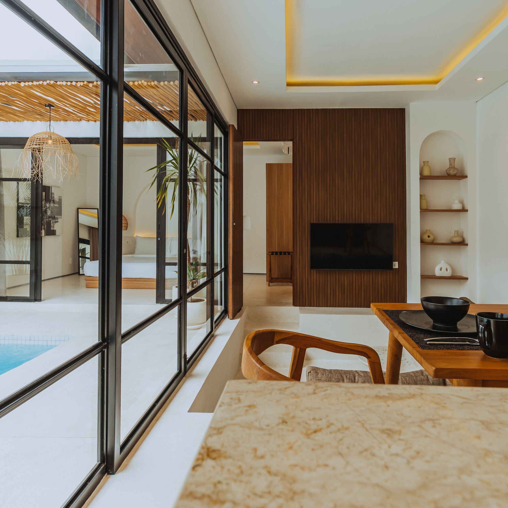 Una sala de estar moderna con un ventanal grande, un comedor con mesa y sillas, un televisor de pantalla plana montado en una pared de paneles de madera, y una piscina visible a través del ventanal.