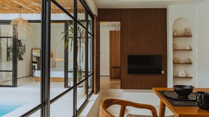 Una sala de estar moderna con un ventanal grande, un comedor con mesa y sillas, un televisor de pantalla plana montado en una pared de paneles de madera, y una piscina visible a través del ventanal.