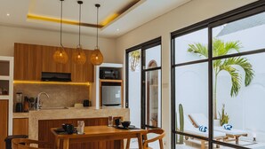 Una cocina moderna con armarios de madera, un comedor con mesa y sillas, y un amplio ventanal con vistas a una zona de piscina con sillones de descanso.