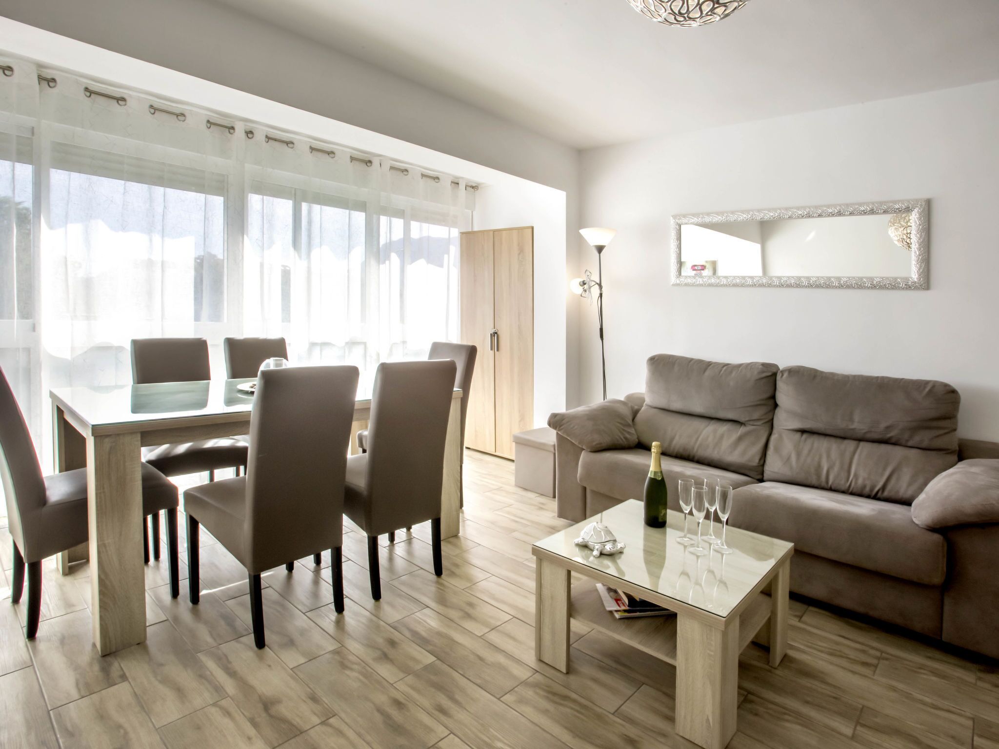 A modern living room with a dining area, a sofa, a coffee table, and a large window with sheer curtains.