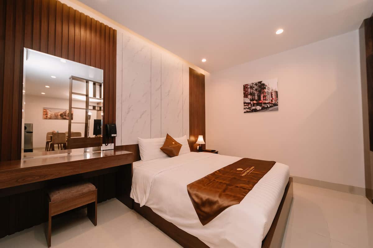 A modern hotel room with a large bed, a wooden desk, a bench, a large mirror, a nightstand with a lamp, and a cityscape artwork on the wall.