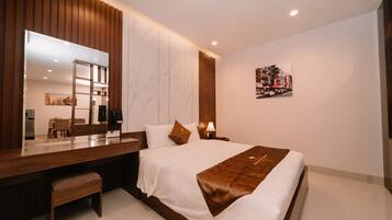 A modern hotel room with a large bed, a wooden desk, a bench, a large mirror, a nightstand with a lamp, and a cityscape artwork on the wall.