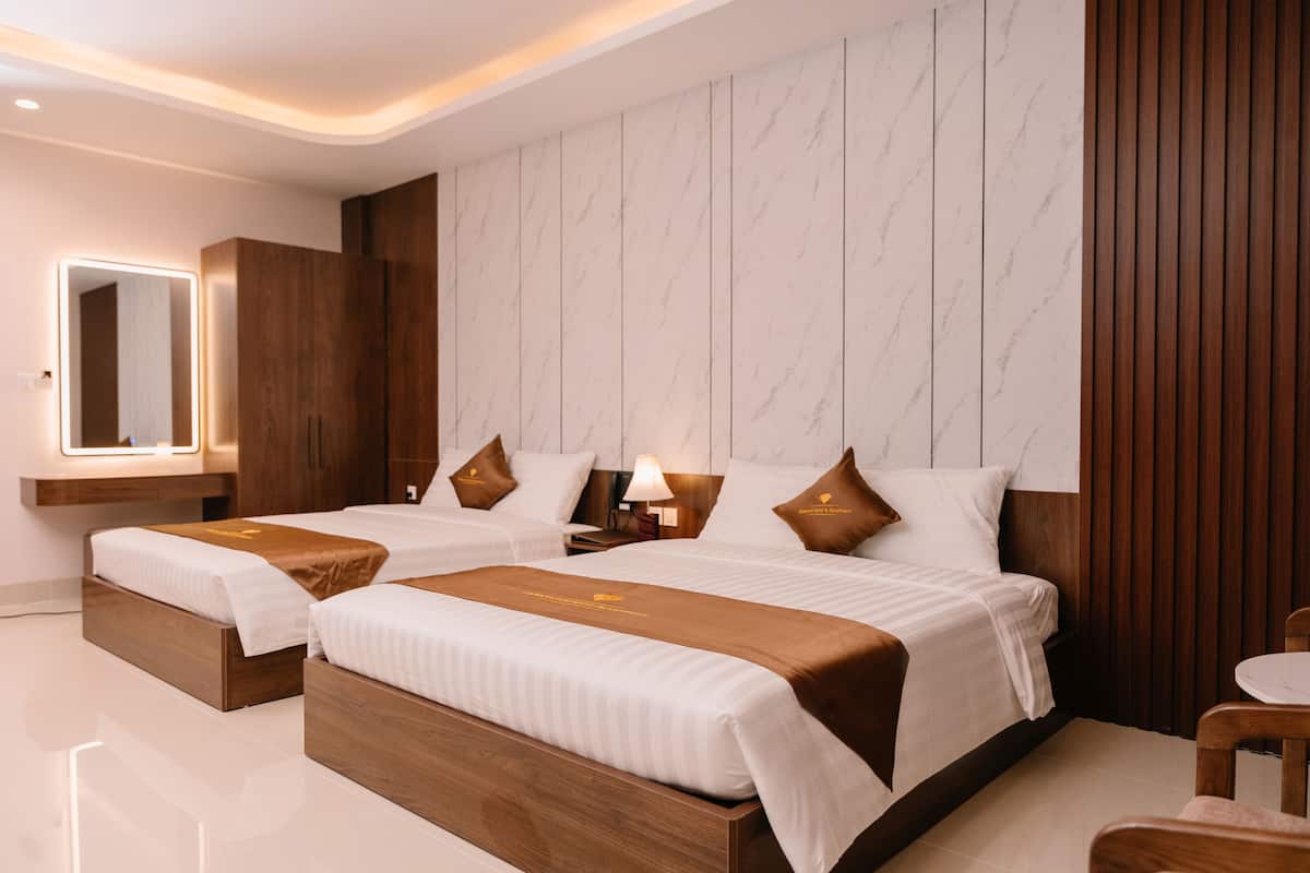 A hotel room with two beds, a wooden wardrobe, a vanity with a mirror, and a bedside table with a lamp.