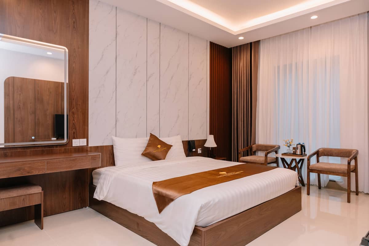 A modern hotel room with a large bed, wooden headboard, a bedside table with a lamp, a small desk, and two chairs.