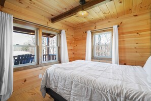 A wooden cabin bedroom with a bed, two windows with curtains, and a view of the outside.