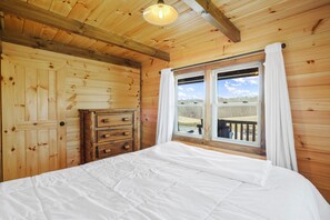 Un dormitorio de cabaña de madera con una cama, un tocador y una ventana con vistas a una terraza y árboles.