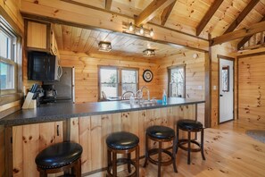 Interior de una cabaña de madera con una zona de cocina que incluye un taburete, un refrigerador y una cafetera.