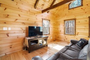 A wooden cabin interior with a television on a wooden cabinet, a leather sofa, and large windows.