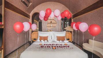 Habitación de hotel con una cama decorada con globos rojos y rosados, un letrero que dice 'Feliz Cumpleaños' y pétalos de rosa.