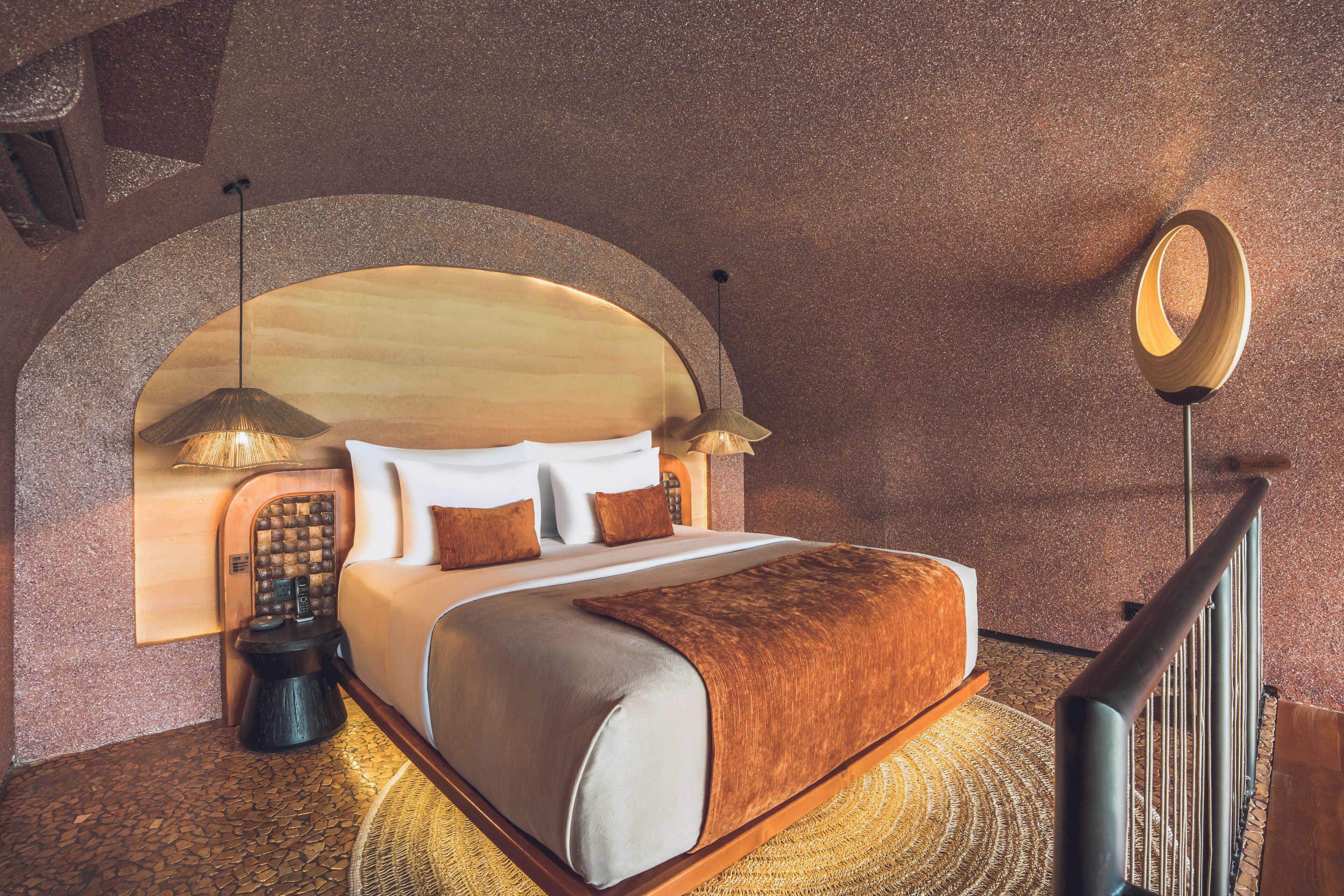 A hotel room with a large bed, a wooden headboard, a round mirror, and a small table with a lamp.