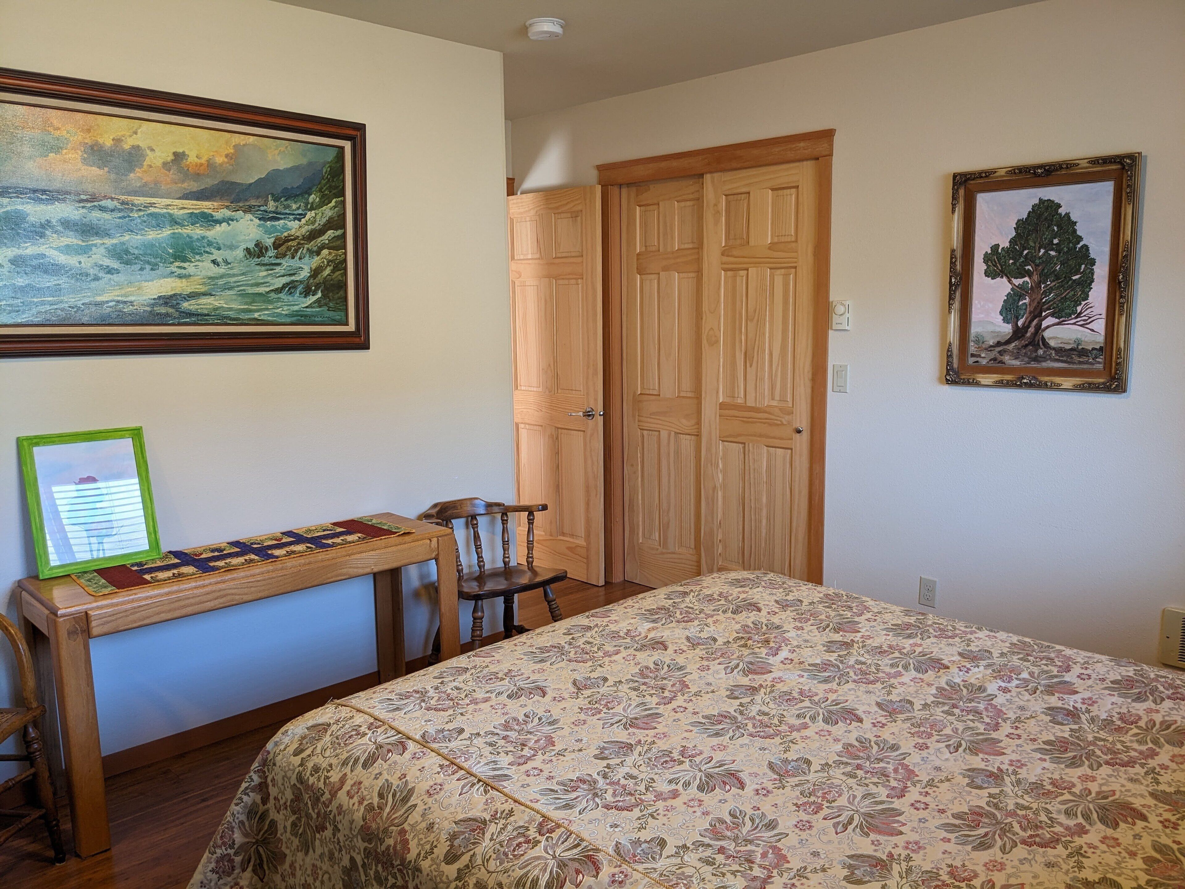 Une chambre à coucher avec un lit, un bureau en bois avec une chaise, un tableau représentant un paysage et une porte.