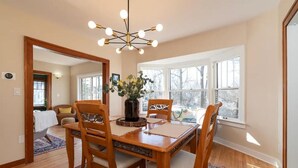 A dining area with a wooden table, four chairs, a chandelier, and large windows. - Historic Craftsman, just 8 minutes from downtown Kalamazoo. (Kalamazoo)
