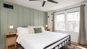 A bedroom with a bed, bedside tables, a ceiling fan, and a window with curtains. - Historic Craftsman, just 8 minutes from downtown Kalamazoo. (Kalamazoo)