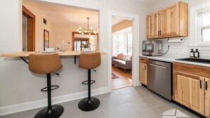 A modern kitchen with wooden cabinets, a stainless steel dishwasher, and a bar area with two stools. - Historic Craftsman, just 8 minutes from downtown Kalamazoo. (Kalamazoo)