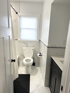 A bathroom with a toilet, a window with blinds, a black floor mat, and a white sink.
