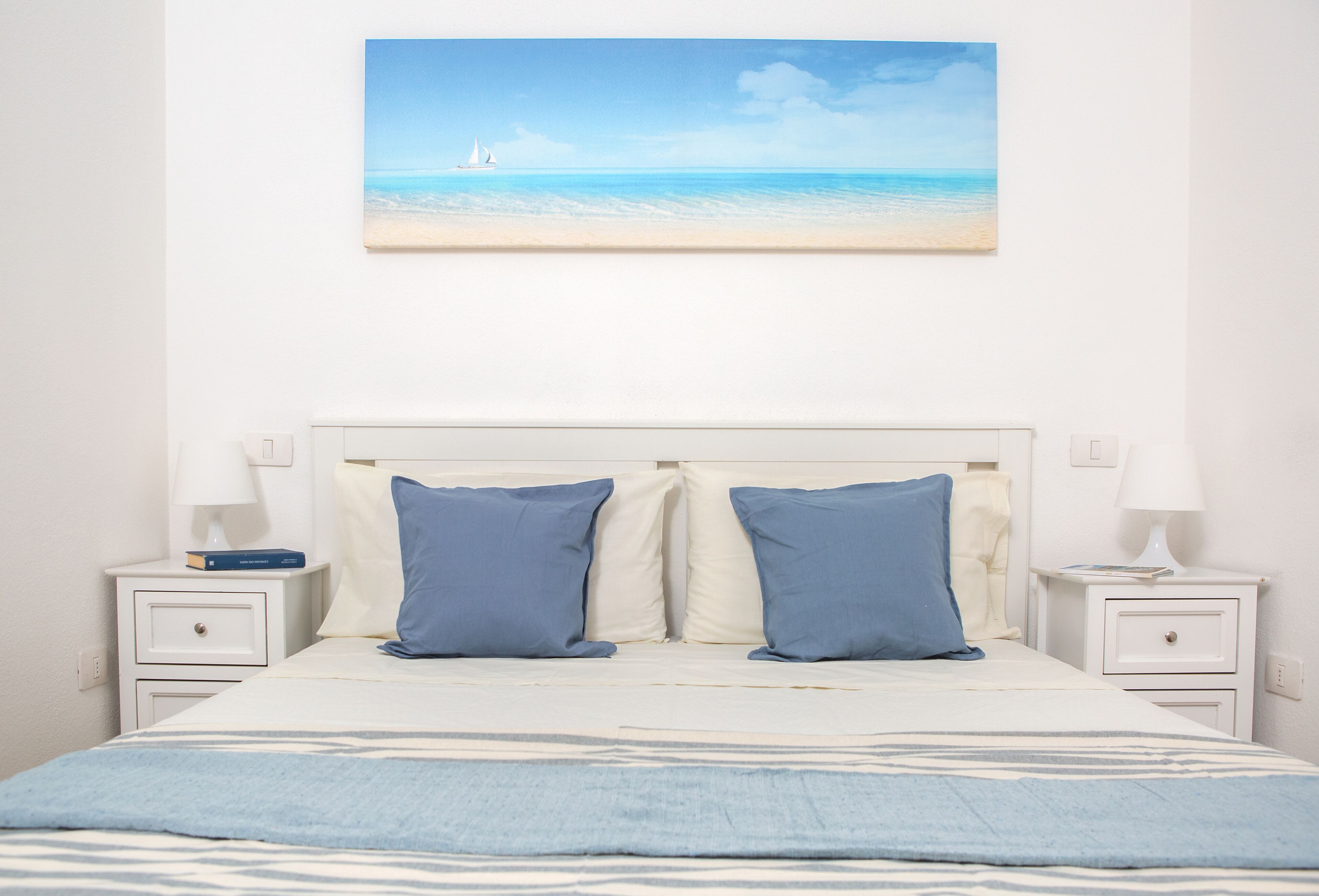 A bedroom with a bed, two pillows, two bedside tables, and a painting of a beach scene.