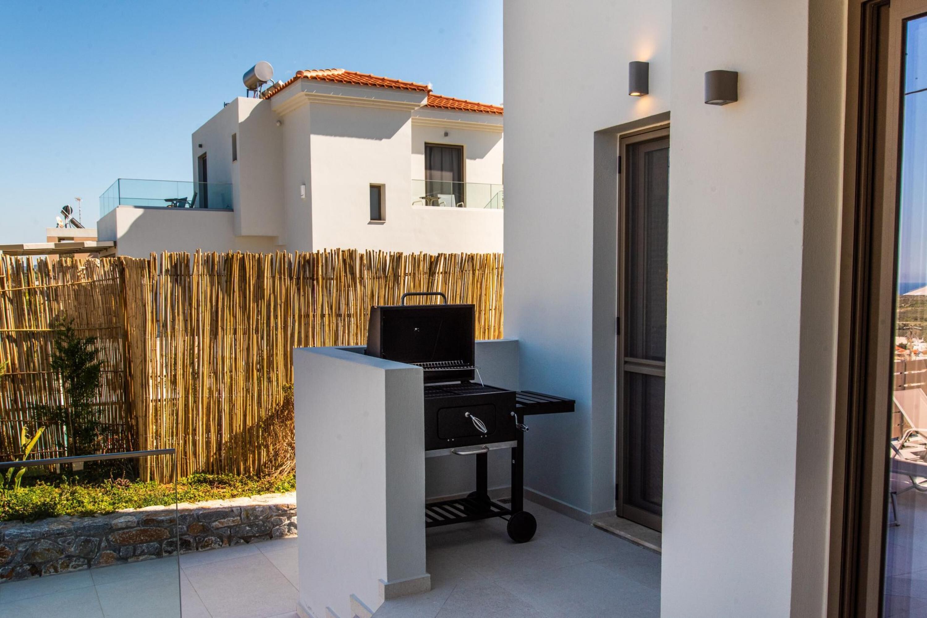 A modern outdoor kitchen area with a grill, a bamboo fence, and a view of a white building with a red roof.