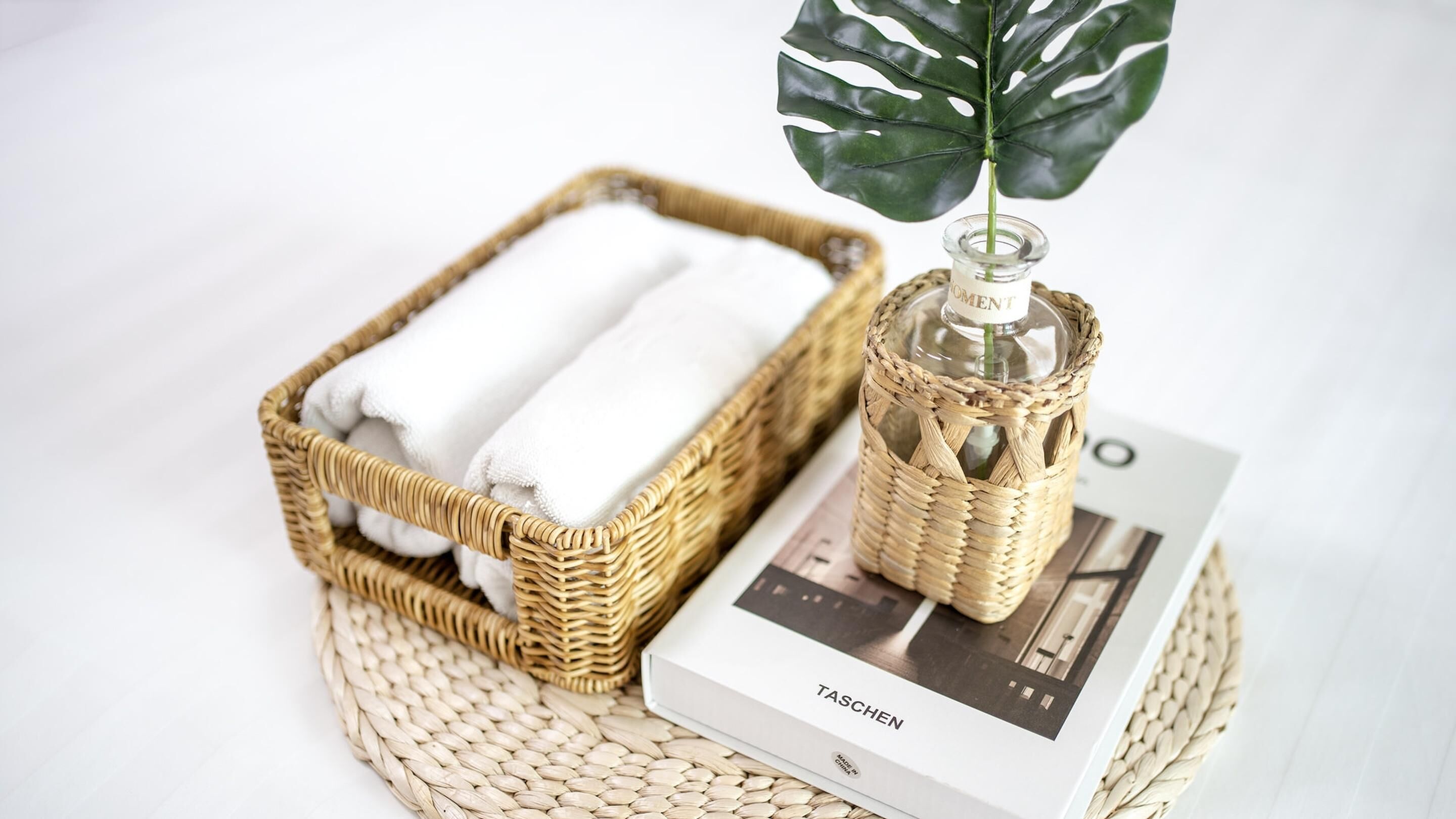 A wicker basket with folded towels, a glass bottle with a label, and a book on a woven mat.