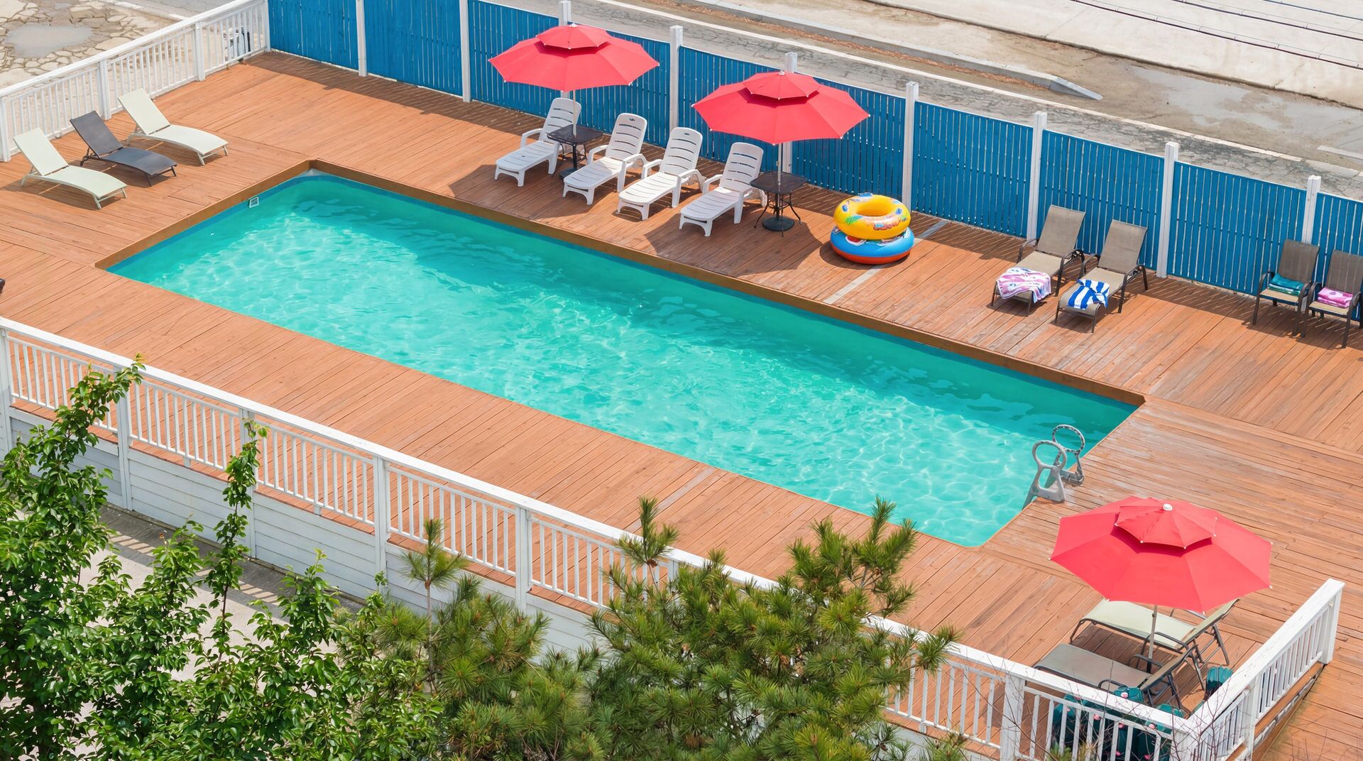 A rectangular pool with clear blue water, surrounded by a wooden deck. There are white lounge chairs and red umbrellas, and a small inflatable ring float.