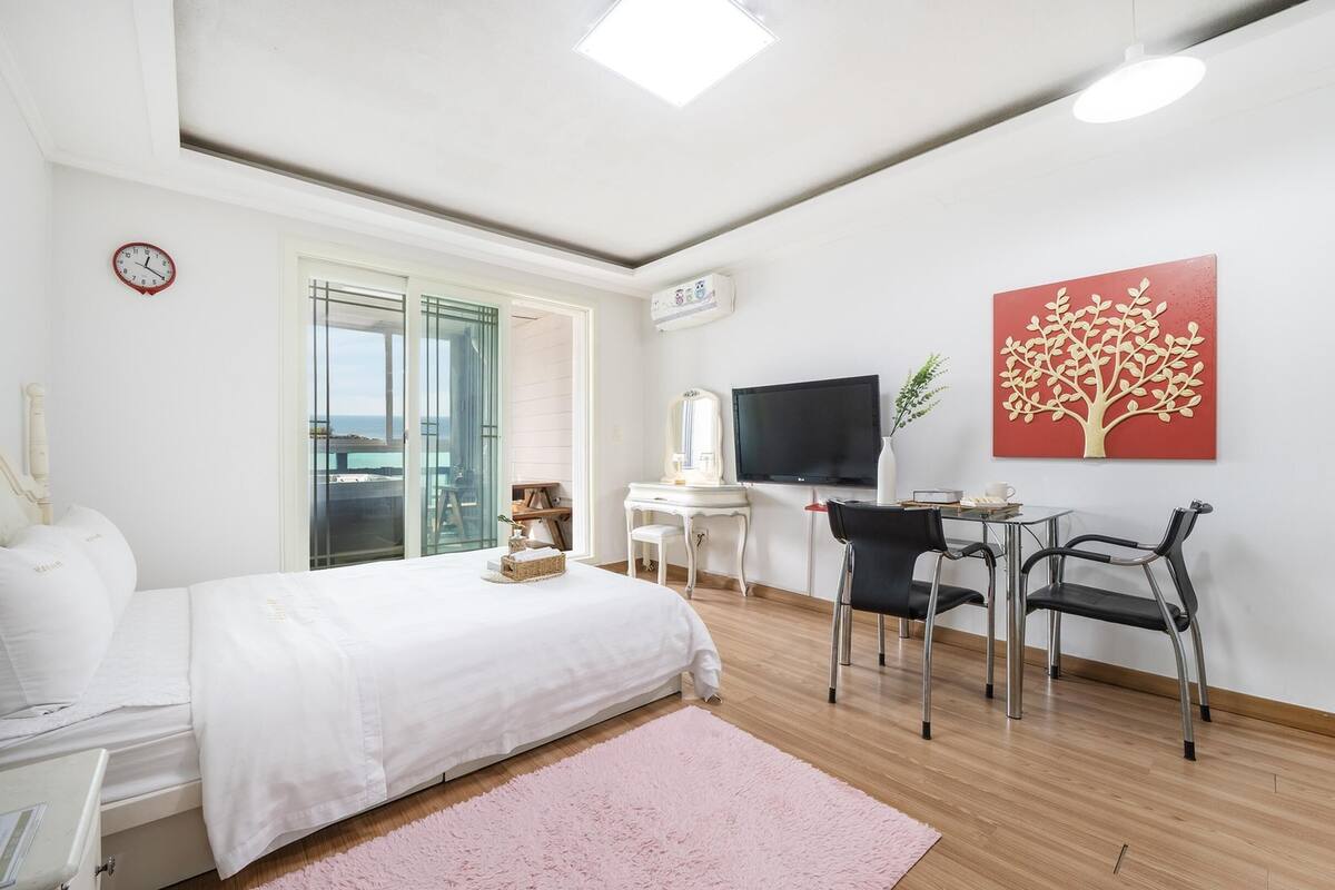 A modern bedroom with a bed, desk, TV, and a view of the sea.