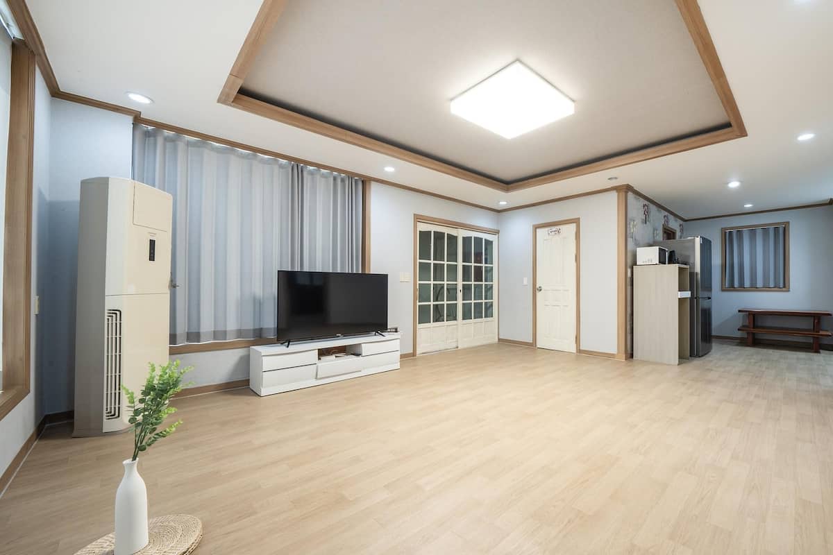 A spacious living room with a flat-screen TV, wooden flooring, a large window with curtains, a built-in wardrobe, and a kitchen area with a refrigerator.