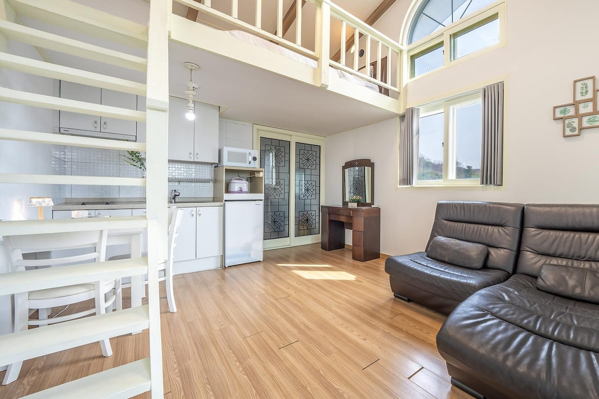 A spacious living area with a high ceiling, a dining table, a sofa, and a kitchen area with a microwave and cabinets.
