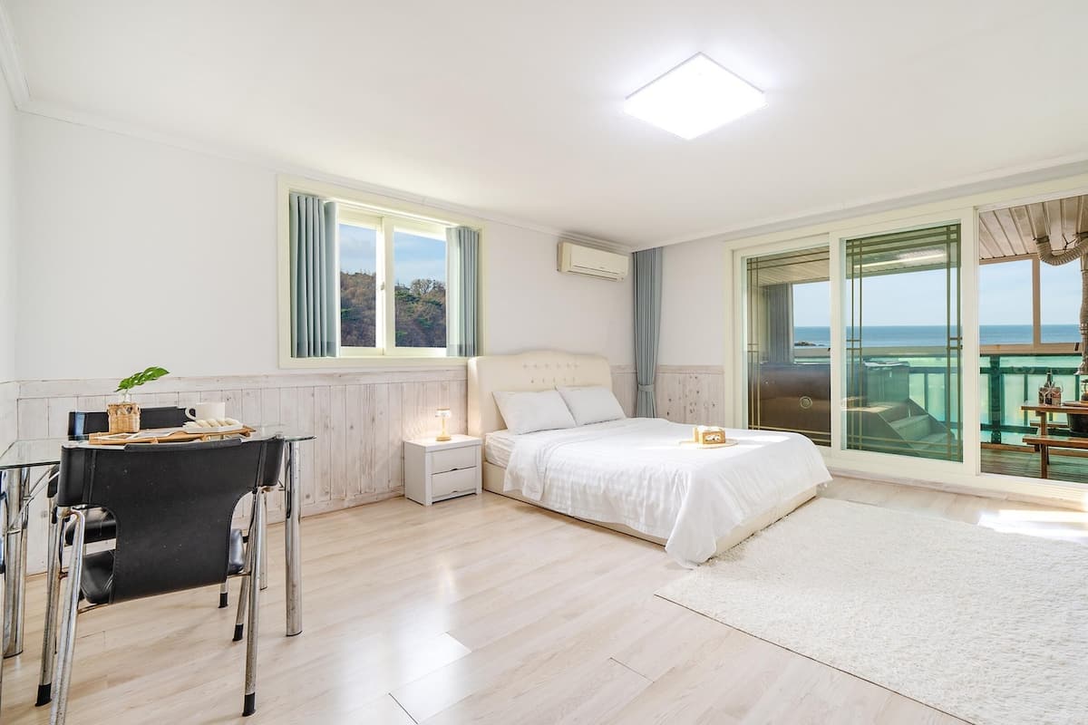 A bedroom with a bed, bedside tables, a desk, and a chair. There is a large window with curtains, a view of the sea, and a balcony with a table and chairs.