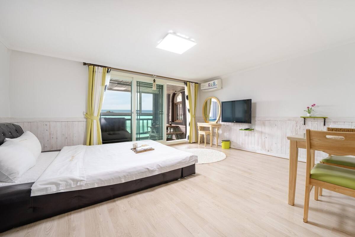 A bedroom with a bed, a desk, a chair, a television, and a balcony with curtains.
