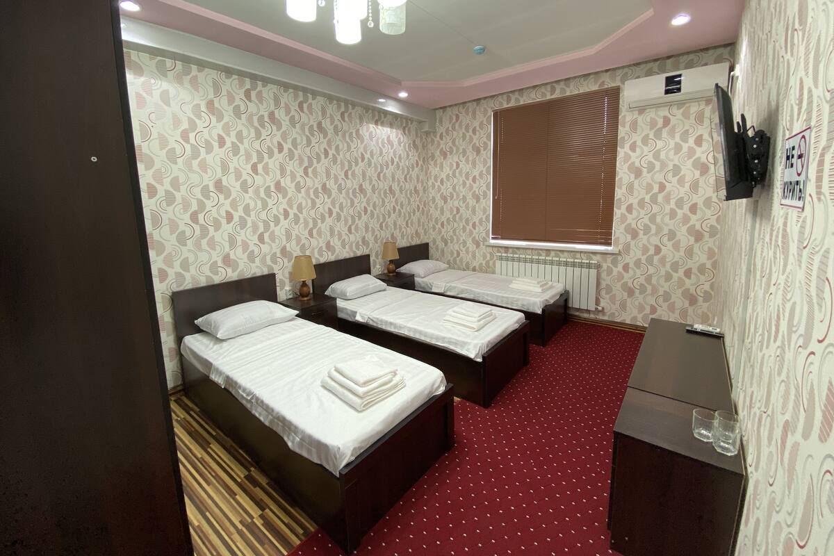 A hotel room with three beds, a red carpet, patterned wallpaper, a ceiling light fixture, and a radiator.