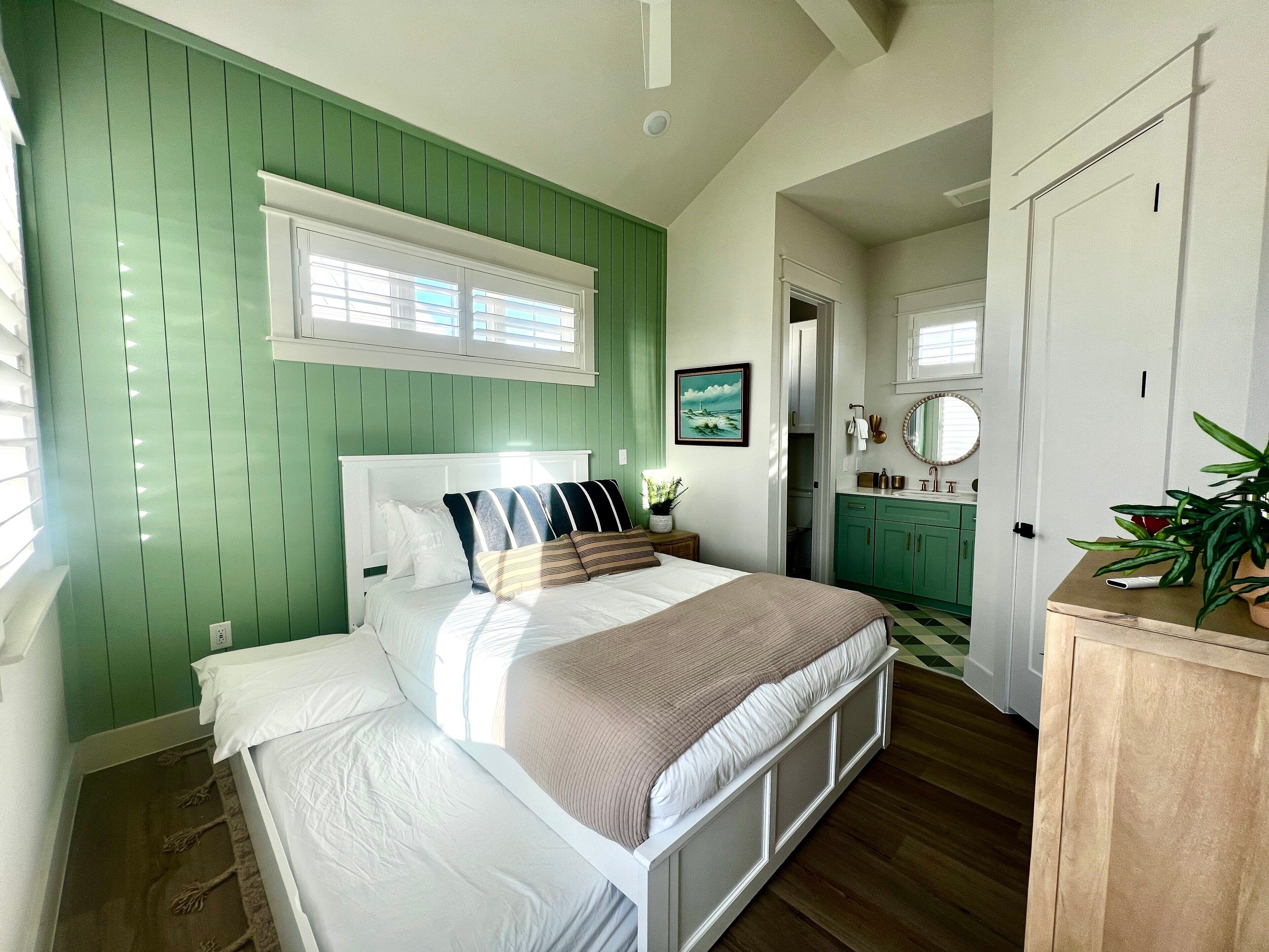 A bedroom with a bed, green walls, a window, a bathroom with green cabinets, and a mirror.