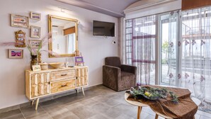 A room with a wooden cabinet, a brown armchair, a television mounted on the wall, and a large window with sheer curtains. - Magical View Studios 1 Asproneri, Kamena Vourla (Kamena Vourla)