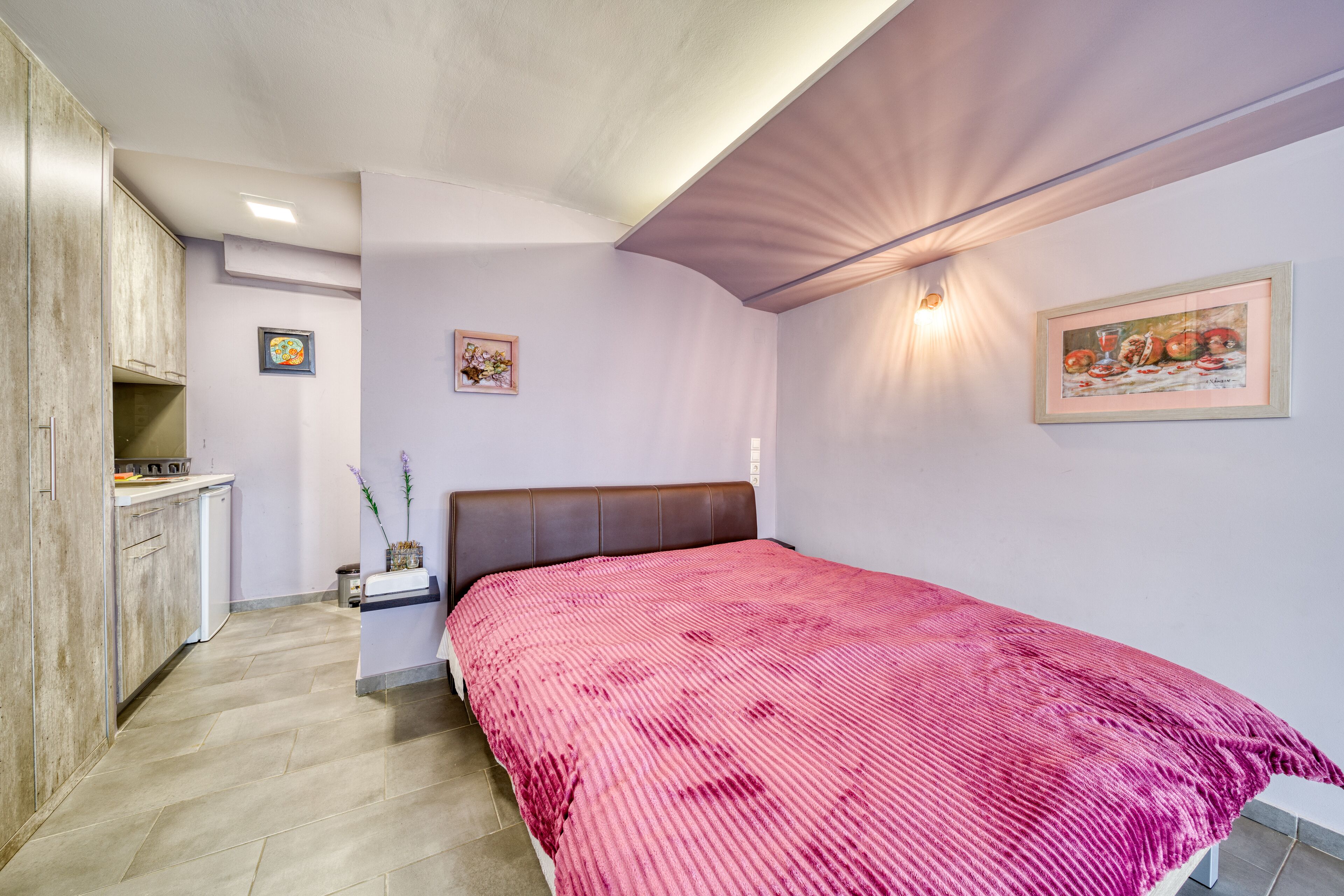 A bedroom with a bed, a nightstand, a wall-mounted shelf, a framed picture, and a kitchenette area.