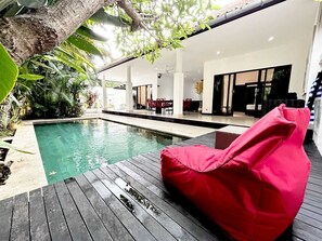 Uma área de piscina moderna ao ar livre, com deck de madeira, uma poltrona de fibra e área de refeições com cadeiras vermelhas.