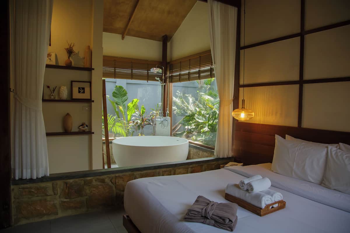Une chambre d’hôtel avec un grand lit, une baignoire ronde et une vue sur une végétation luxuriante par la fenêtre.