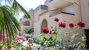 Un immeuble à plusieurs étages doté de balcons, avec un palmier et des fleurs roses et rouges au premier plan.