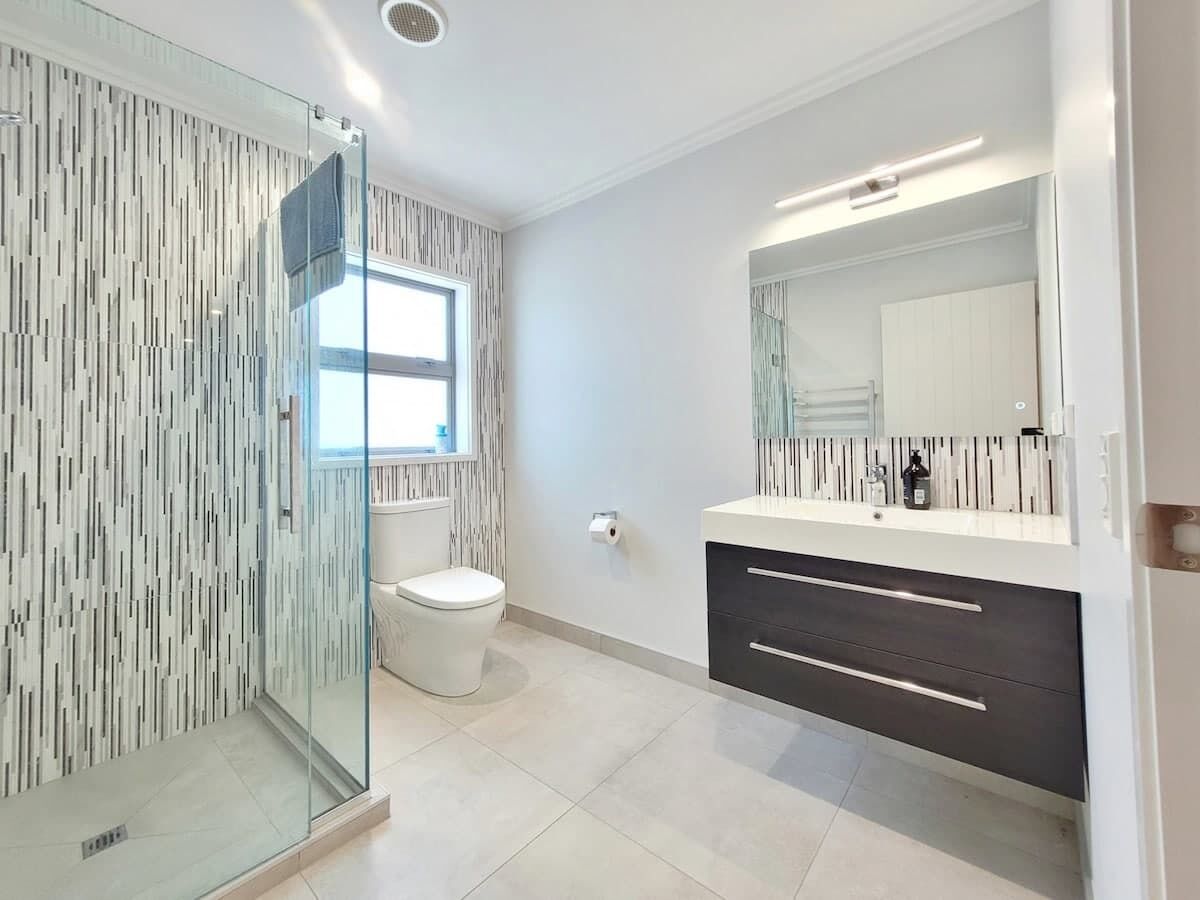 Une salle de bain moderne avec une douche à parois de verre, des toilettes blanches et un meuble-lavabo foncé doté d’un miroir.