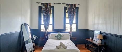 Un dormitorio con cama, ventilador de techo, ventana con cortinas, mesita de noche con lámpara y espejo.
