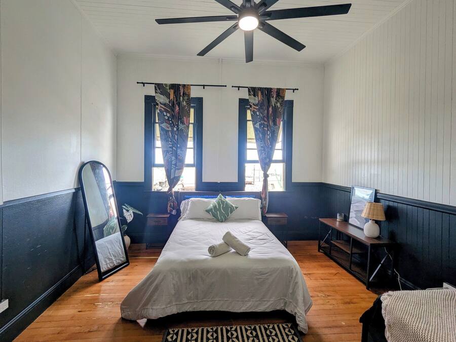 Un dormitorio con cama, ventilador de techo, ventana con cortinas, mesita de noche con lámpara y espejo.