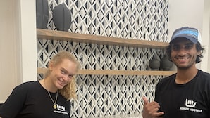 Dos personas cocinando en una plancha, con una pared decorada y utensilios de cocina al fondo.
