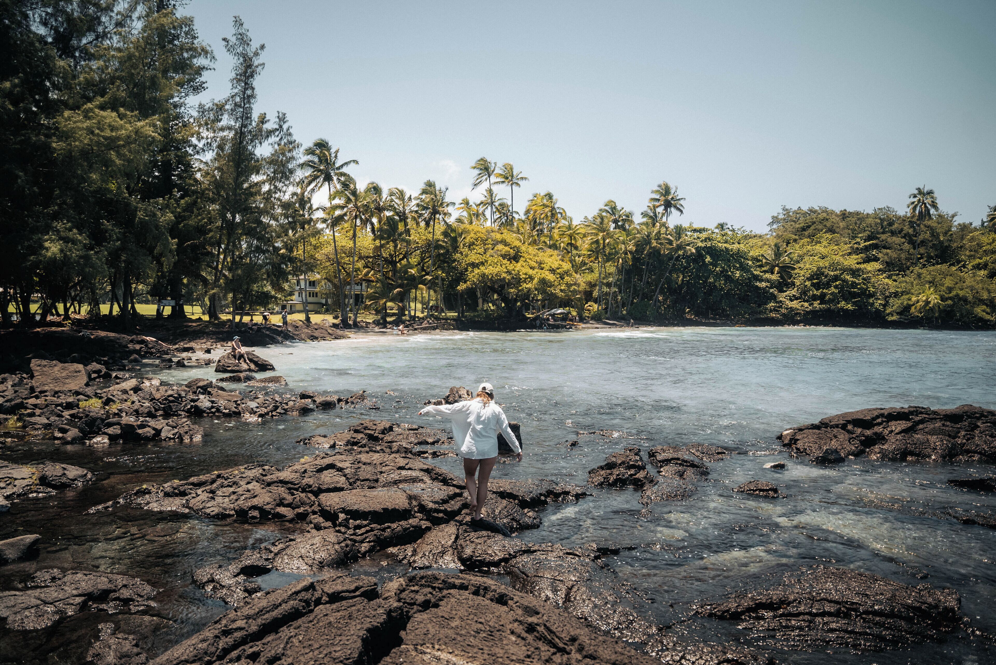 A rocky shoreline with a person standing on rocks, palm trees in the background, and clear water.