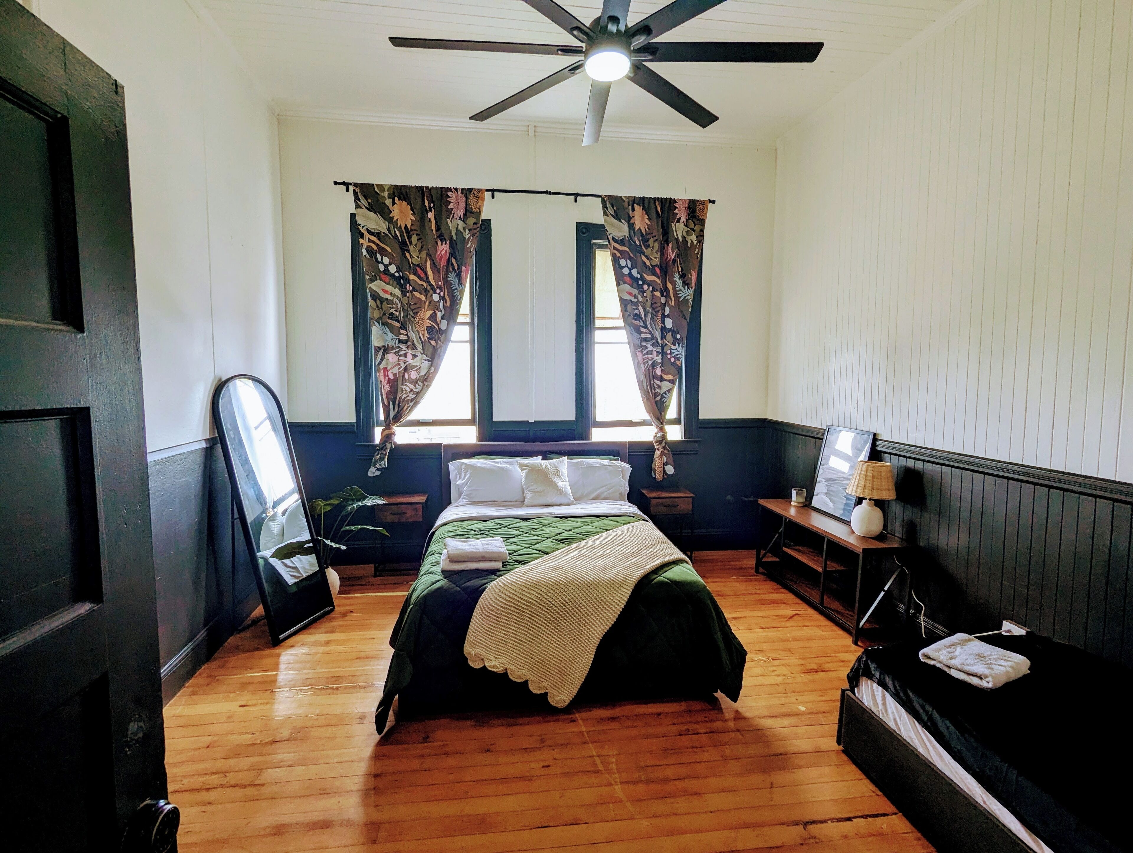 Un dormitorio con cama, ventilador de techo, ventana con cortinas, mesita de noche con lámpara y suelo de madera.
