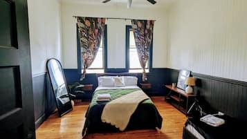 Un dormitorio con cama, ventilador de techo, ventana con cortinas, mesita de noche con lámpara y suelo de madera.