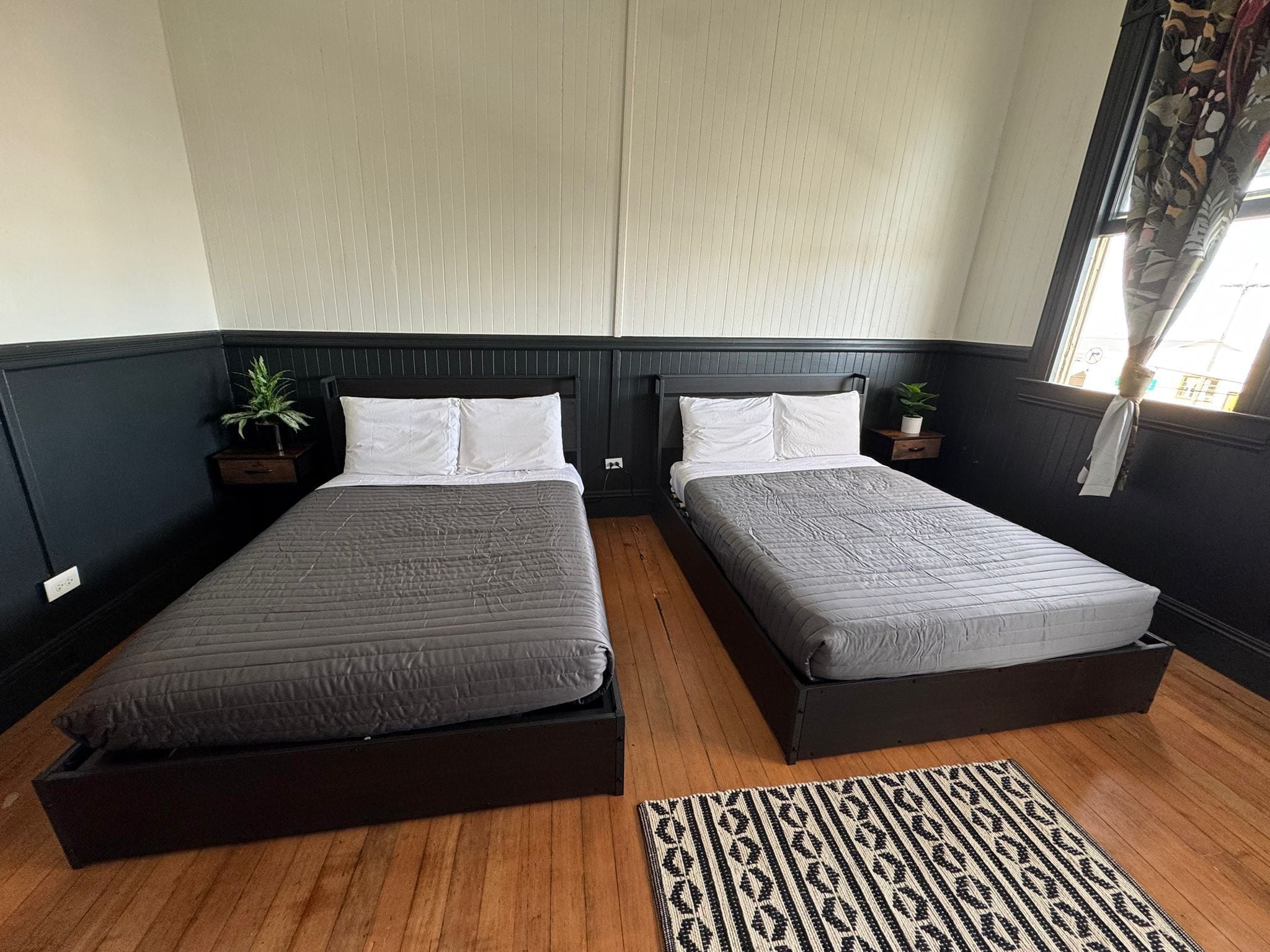 A room with two single beds, a wooden floor, a patterned rug, and a window with curtains.