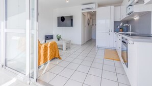 A modern kitchen with white cabinets, a built-in oven, and a sink. There is a dining area with a table and chairs, and a TV mounted on the wall. - Grand Bleu by Interhome (Le Barcarès)