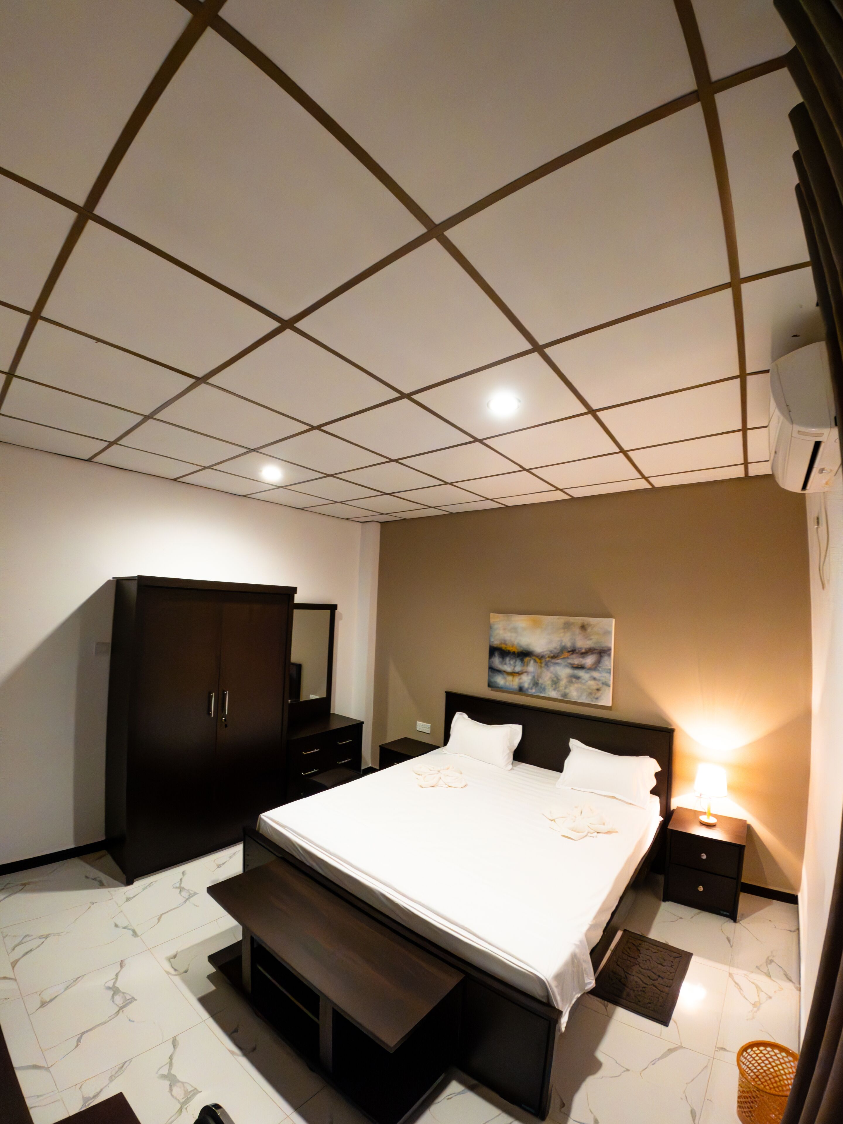A bedroom with a large bed, a wardrobe, a bedside table, a lamp, and a ceiling with recessed lighting.