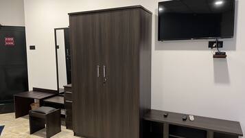 Una habitación con un armario de madera oscura, un pequeño escritorio con cajones, un televisor montado en la pared y un banco con un control remoto encima.