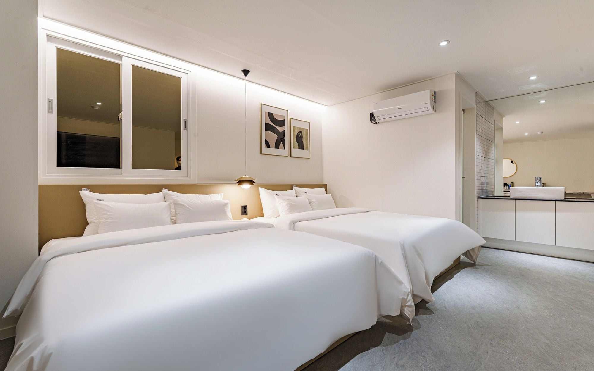 Camera d'albergo con due letti, un'ampia finestra, un condizionatore e un bagno parzialmente visibile.